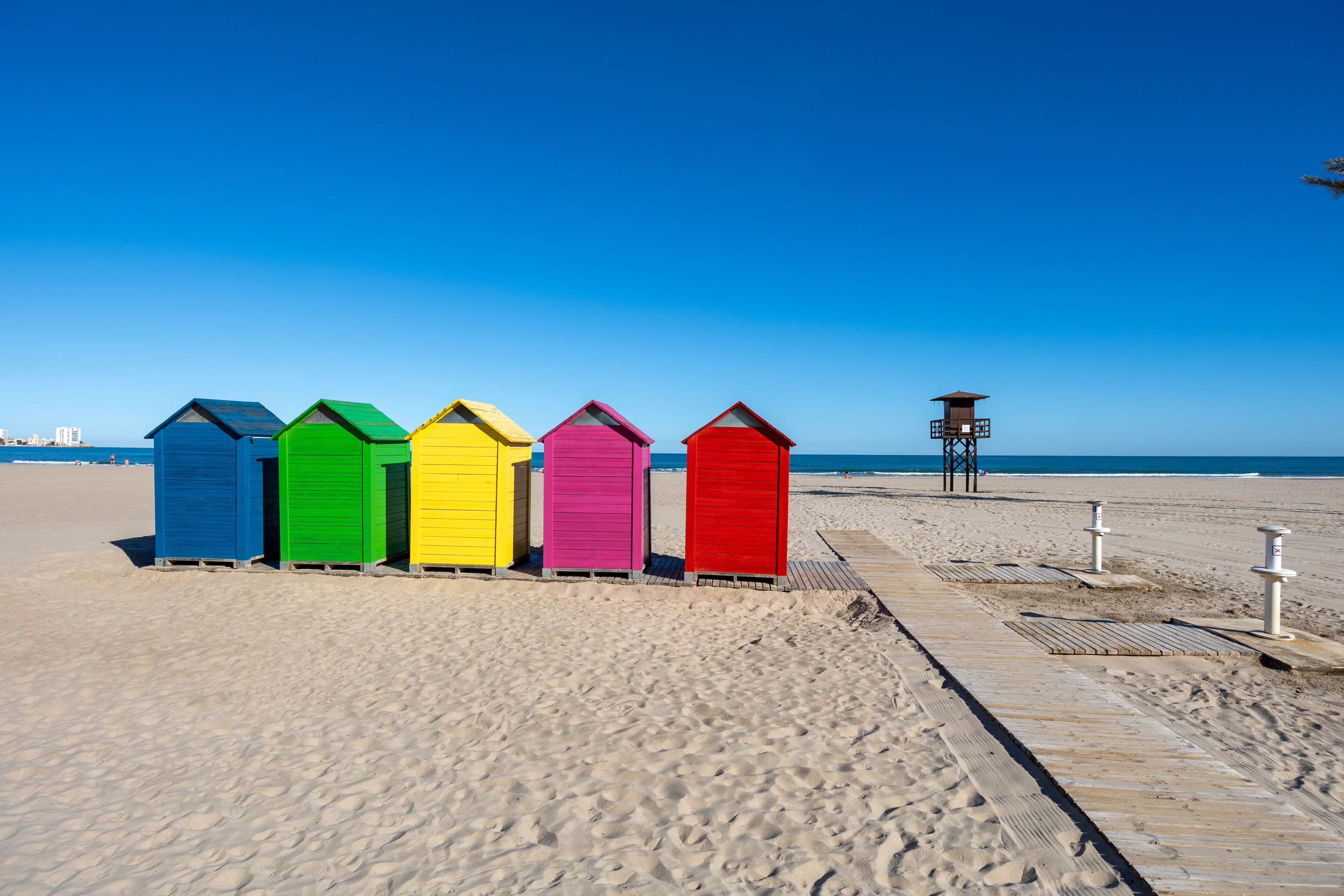 A row of colorful beach huts on a sandy beach with a clear blue sky.