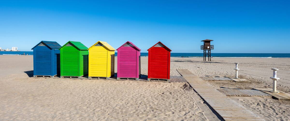 Una hilera de cabañas de playa coloridas en una playa arenosa con un cielo azul despejado.