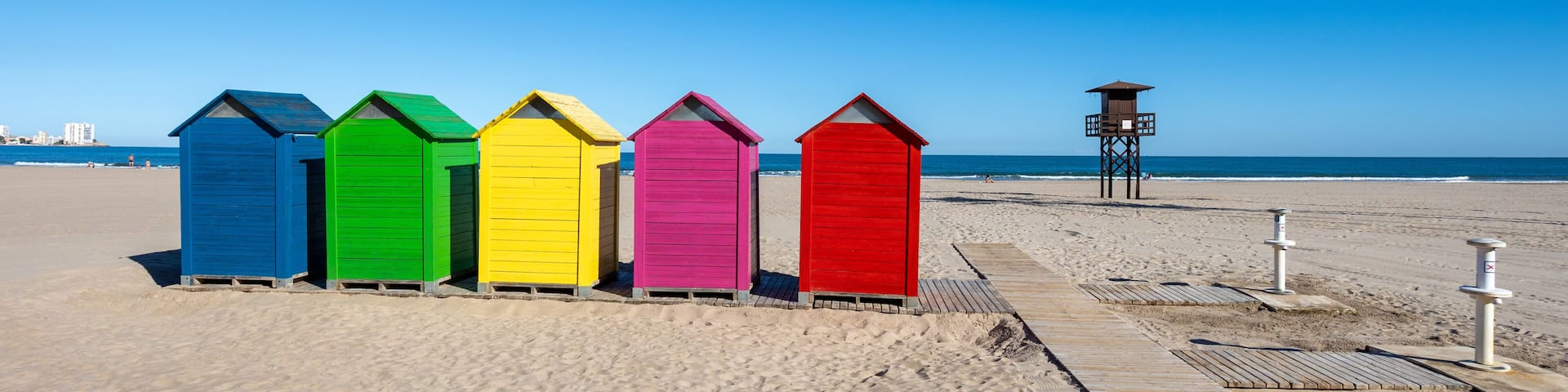 A row of colorful beach huts on a sandy beach with a clear blue sky.
