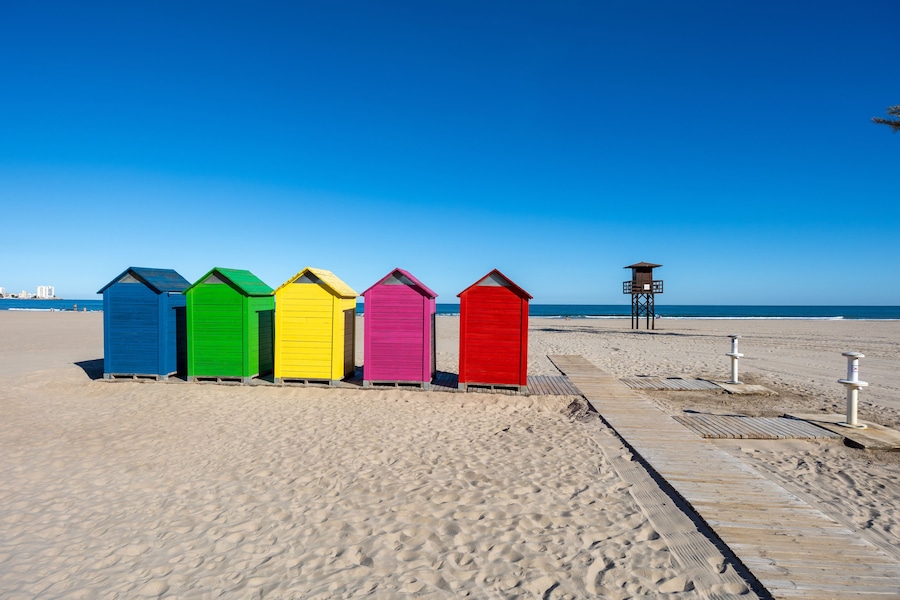 A row of colorful beach huts on a sandy beach with a clear blue sky.