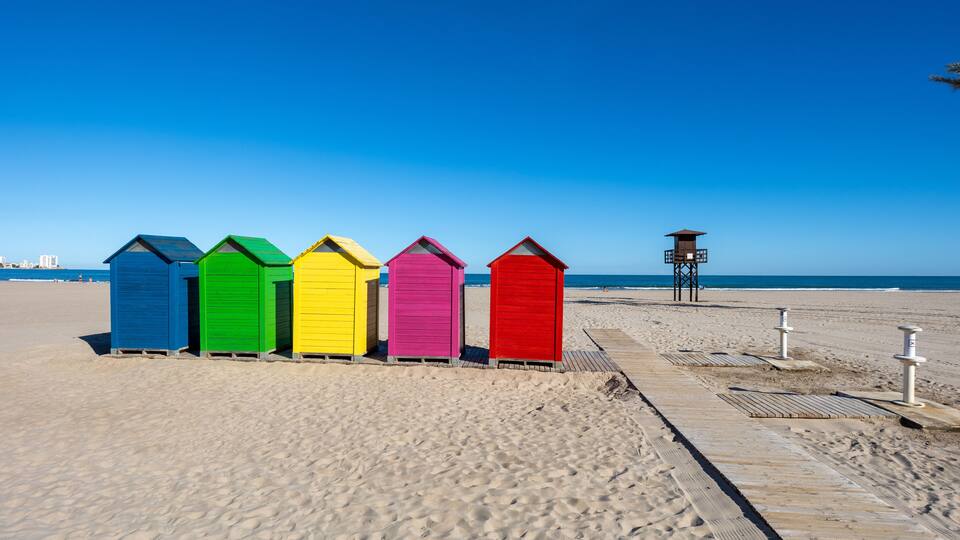 A row of colorful beach huts on a sandy beach with a clear blue sky.