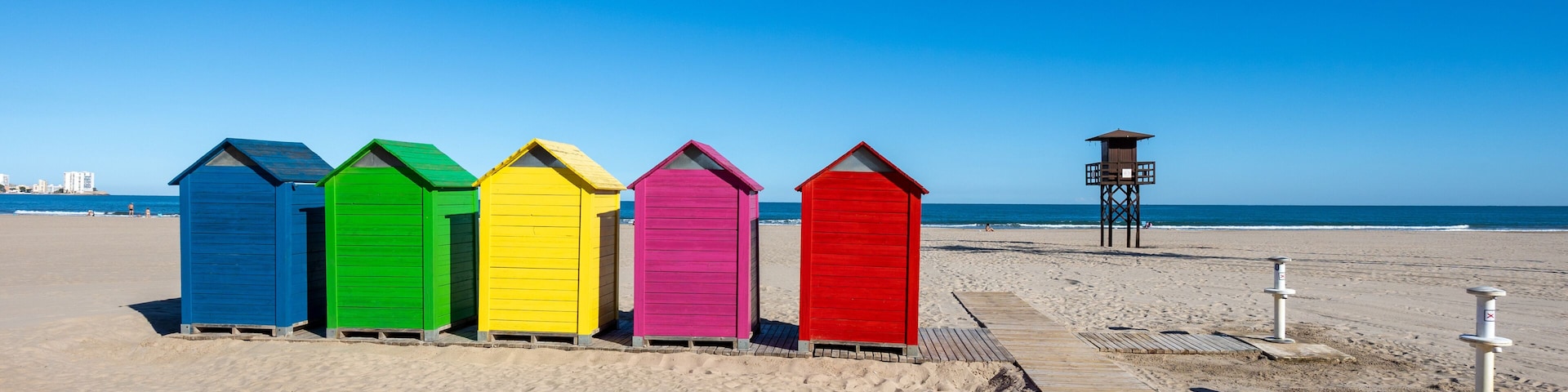 Uma fileira de cabanas coloridas numa praia arenosa, sob um céu azul e límpido.