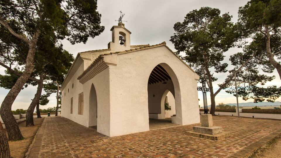 A small white church with a bell tower and arched entrance, surrounded by trees and a paved walkway.