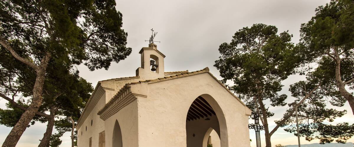 A small white church with a bell tower and arched entrance, surrounded by trees and a paved walkway.