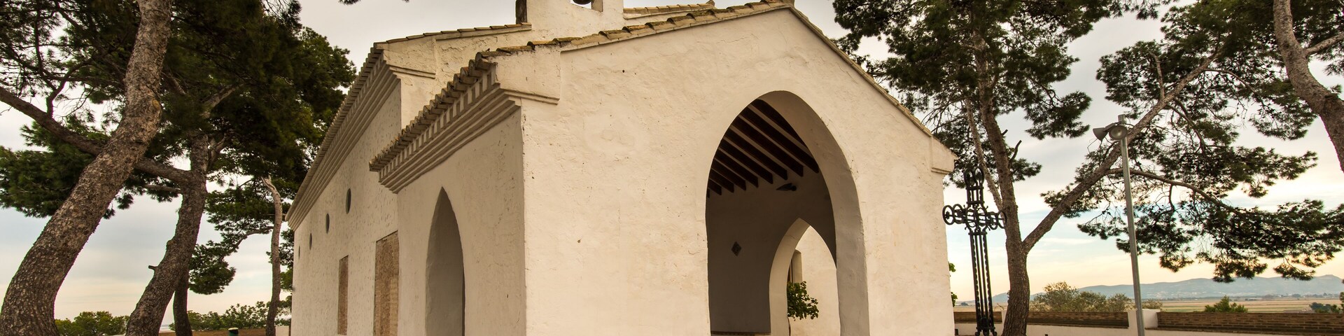A small white church with a bell tower and arched entrance, surrounded by trees and a paved walkway.