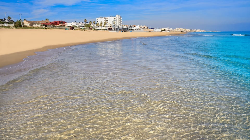 A beach with clear shallow waters, a sandy shore, and buildings in the background.