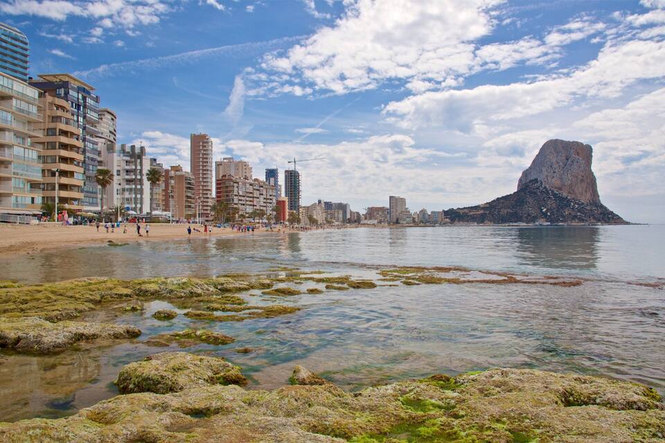 A coastal cityscape with a prominent rock formation, sandy beach, and modern buildings.