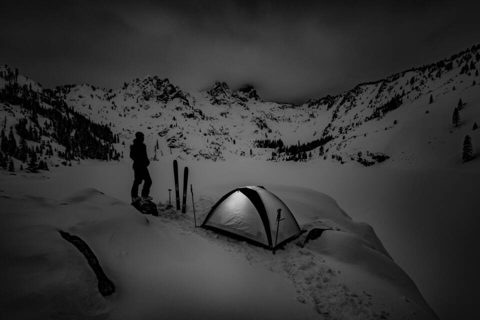 Une personne se tient sur une montagne enneigée, avec une tente et des skis à proximité.