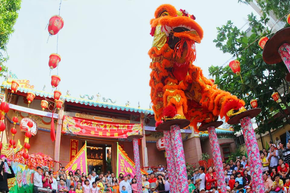 A traditional lion dance performance with a large orange lion costume in front of a building with Chinese decorations and a crowd of spectators.