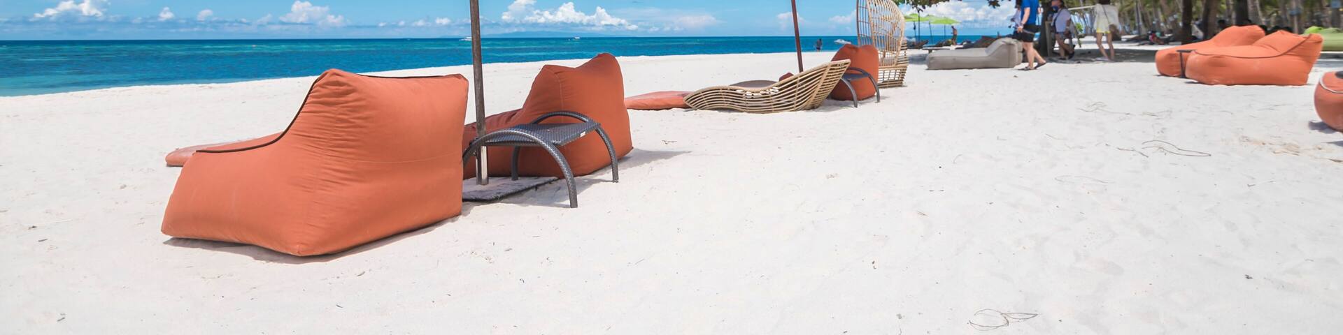 A beach with orange lounge chairs and umbrellas, palm trees, and the ocean in the background.