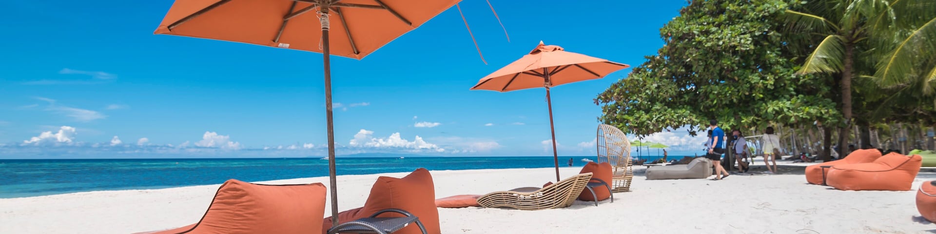 A beach with orange lounge chairs and umbrellas, palm trees, and the ocean in the background.
