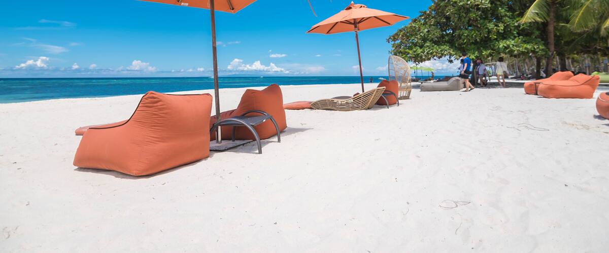 A beach with orange lounge chairs and umbrellas, palm trees, and the ocean in the background.