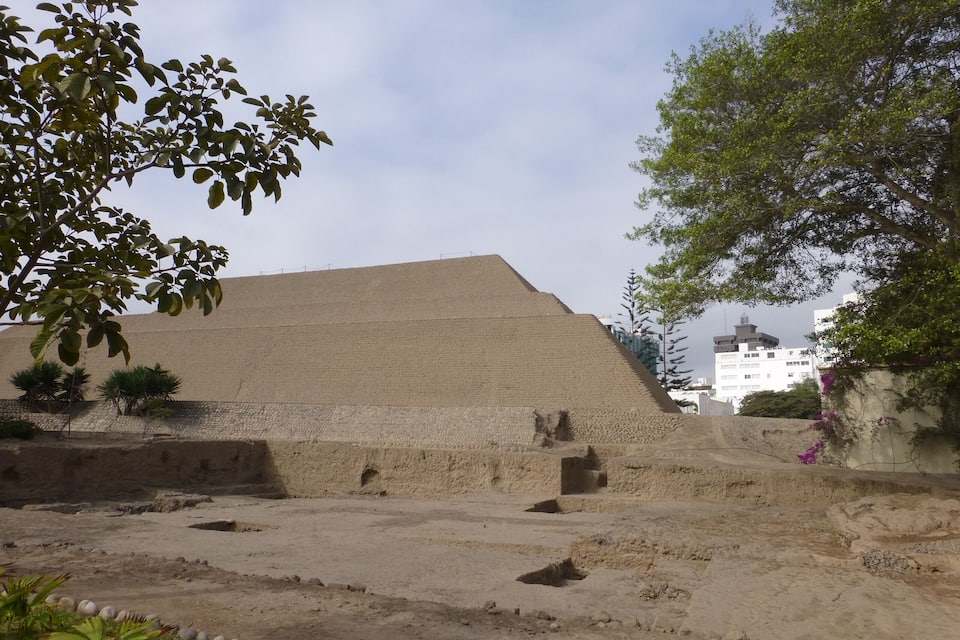 Une grande structure pyramidale à sommet plat, entourée d’une zone sablonneuse parsemée de rochers et où la végétation est clairsemée.