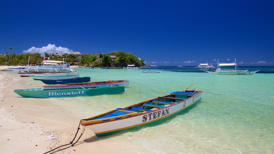 A beach with several colorful boats anchored near the shore, clear turquoise water, and a backdrop of lush greenery and buildings.