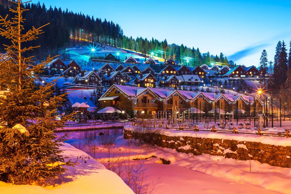 Ein schneebedecktes Dorf mit Holzhäusern und einer Skipiste im Hintergrund.