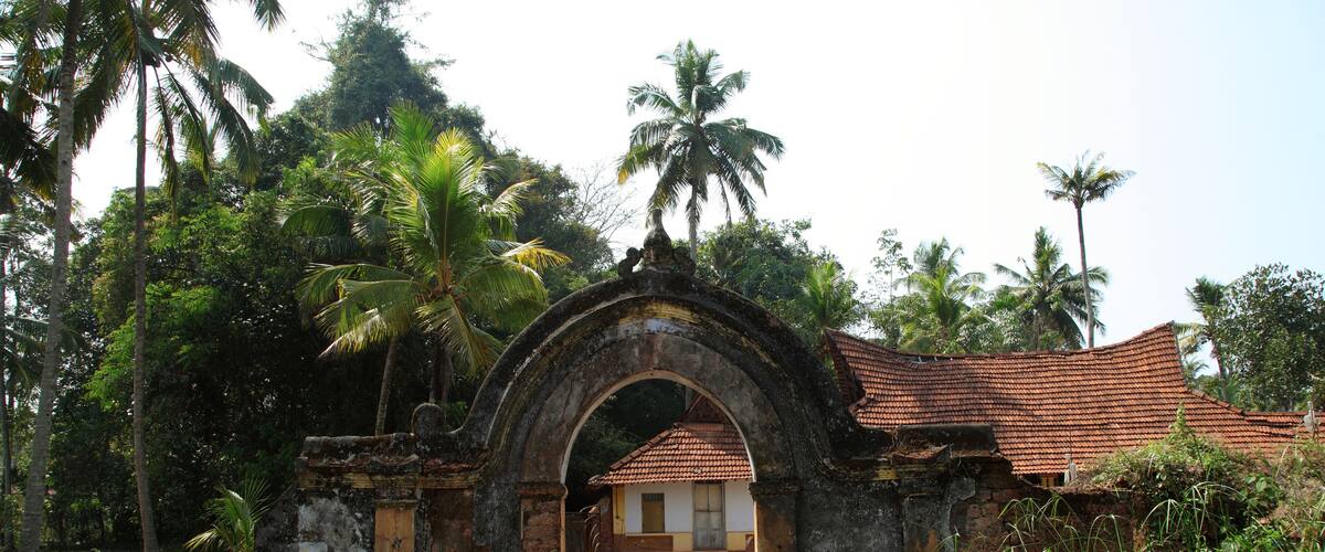 An ancient stone archway leading to a traditional house with a tiled roof, surrounded by lush greenery and palm trees.