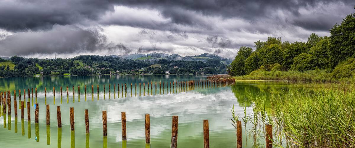 A calm lake with wooden posts in the water, surrounded by lush greenery and mountains under a cloudy sky.