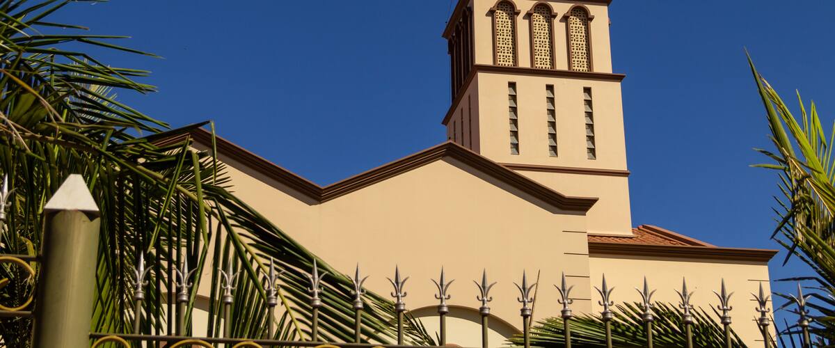 A church with a red-tiled roof and a cross on top, surrounded by palm trees.