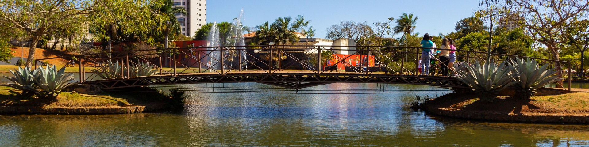 Uma ponte sobre um rio calmo, cercada por árvores e um edifício alto ao fundo.