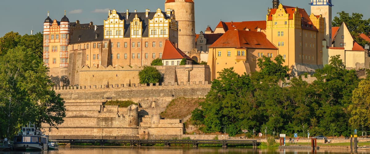 Eine historische Burg mit mehreren Türmen und einem Fluss im Vordergrund.