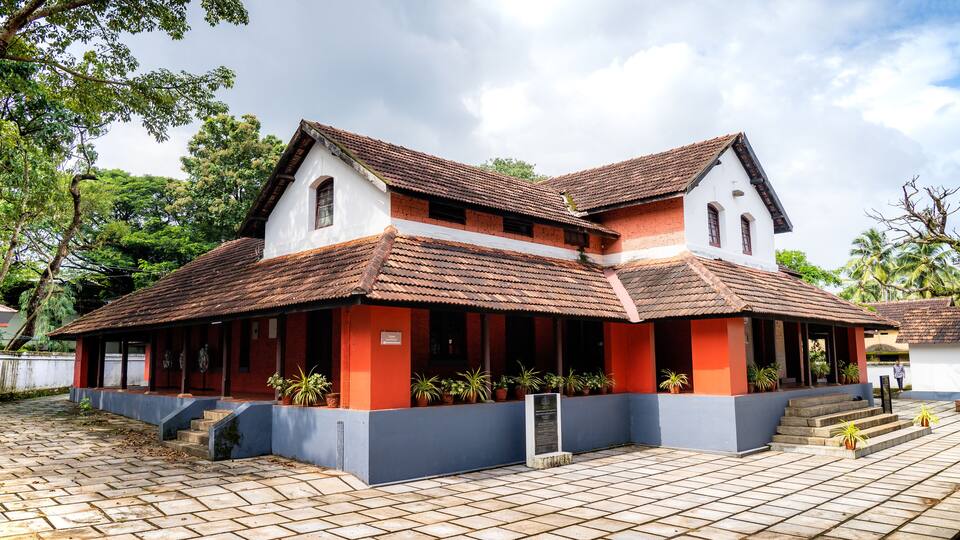 A two-story building with a red roof and white walls, surrounded by potted plants and a paved courtyard.
