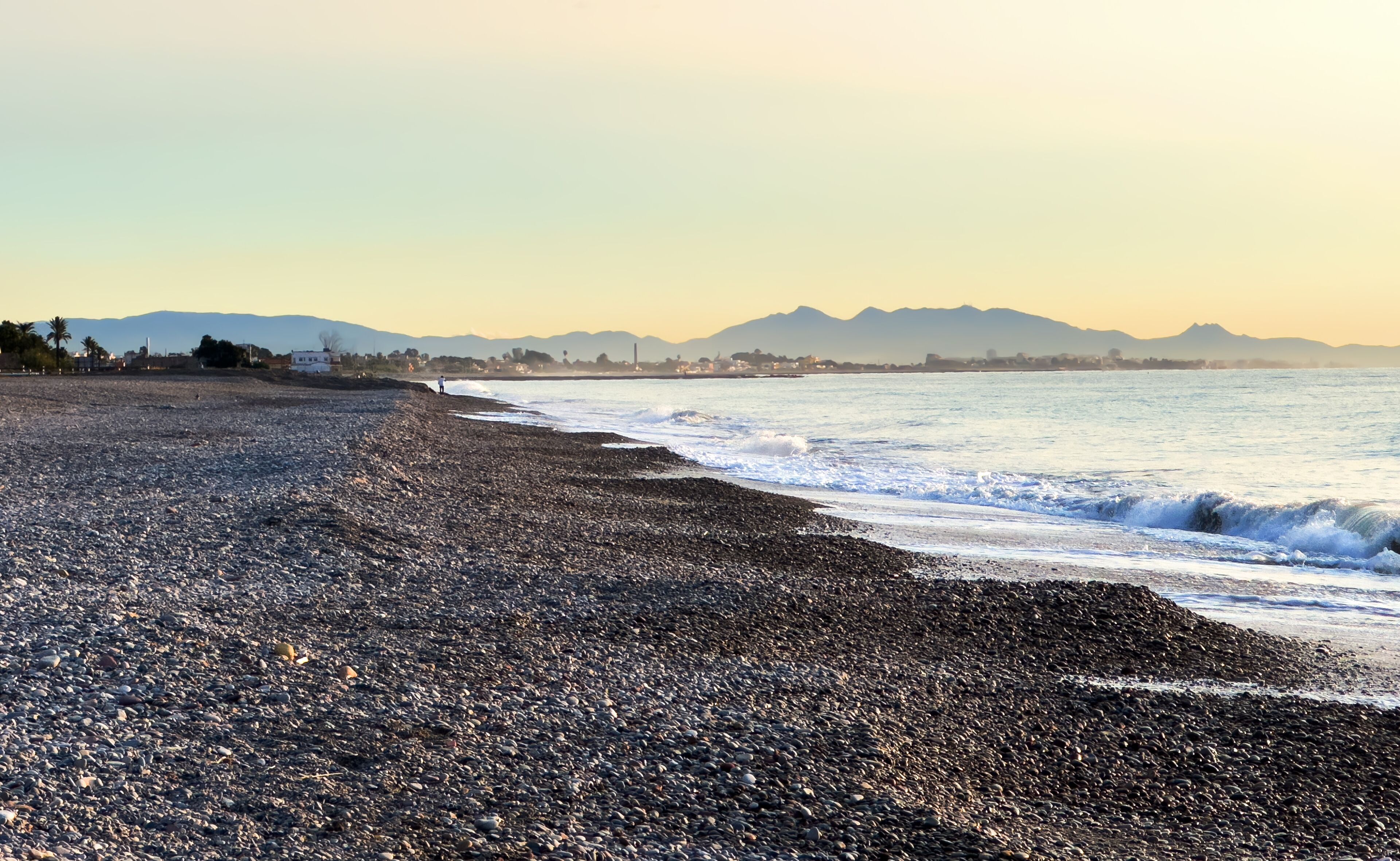 A pebbly beach with waves, mountains, and a clear sky.