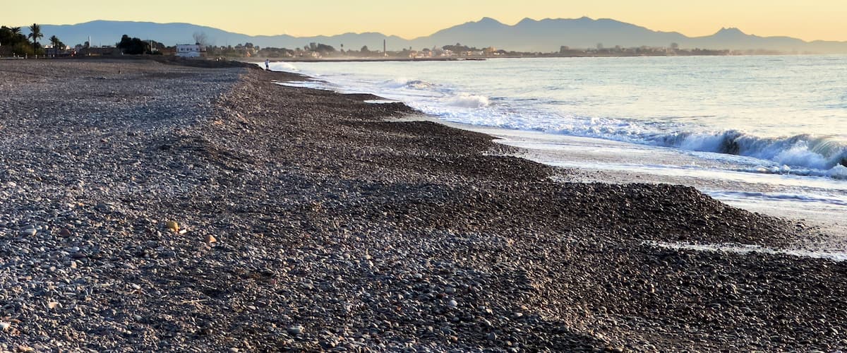 A pebbly beach with waves, mountains, and a clear sky.