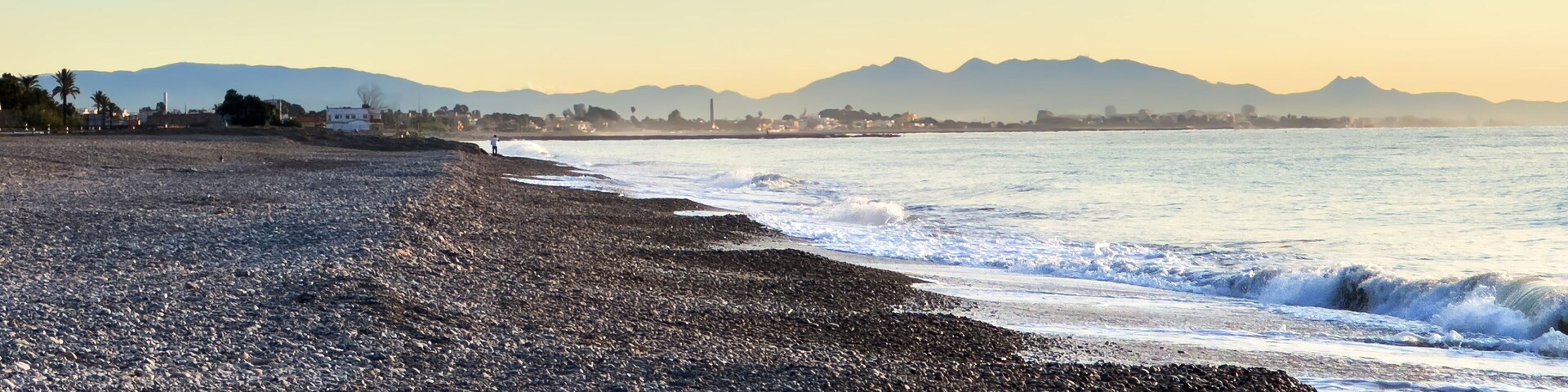 A pebbly beach with waves, mountains, and a clear sky.
