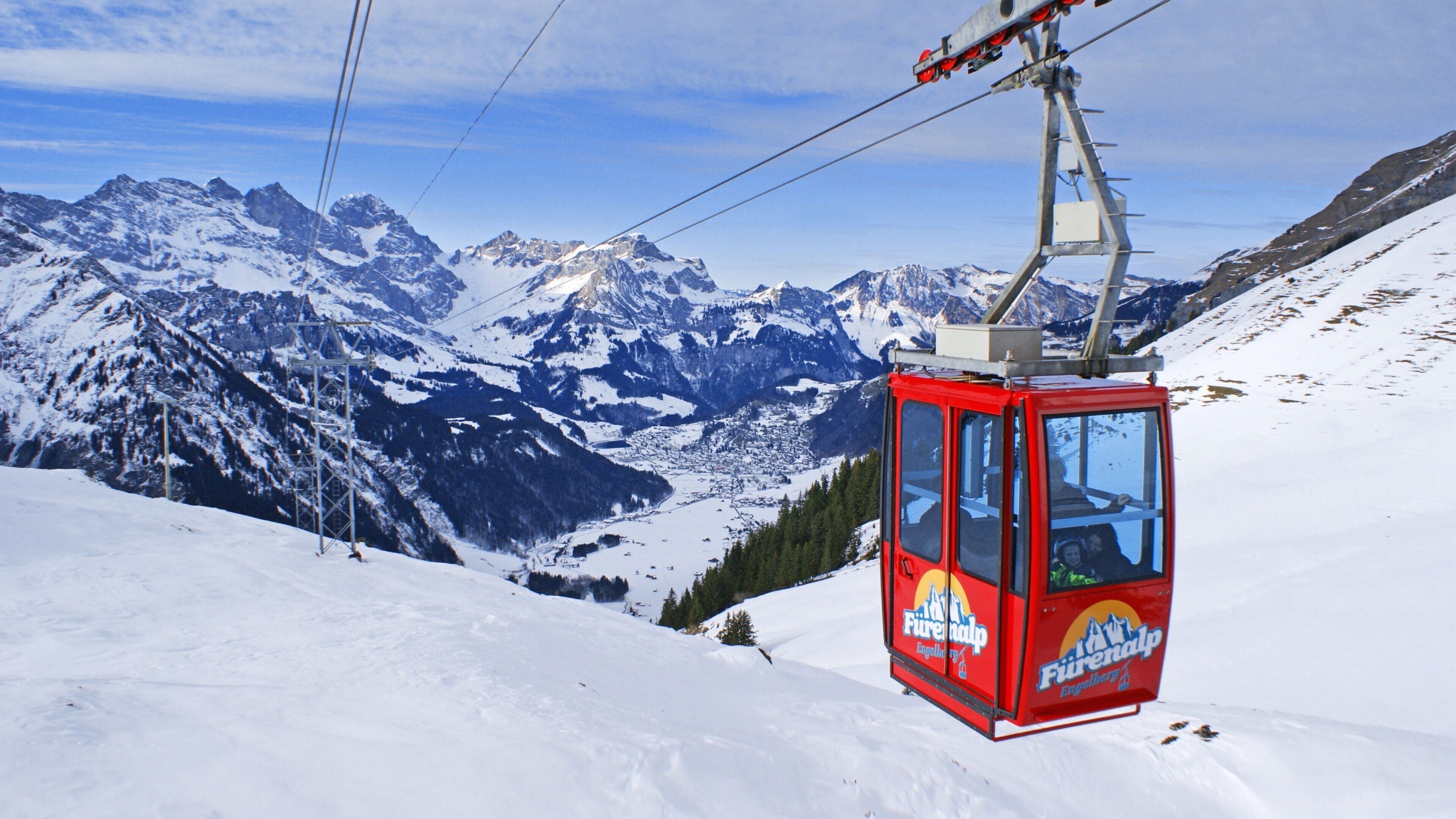 A red cable car with 'Fürenalp Engellbuh' written on it, traveling over a snowy mountain with a backdrop of snow-covered peaks and a clear sky.