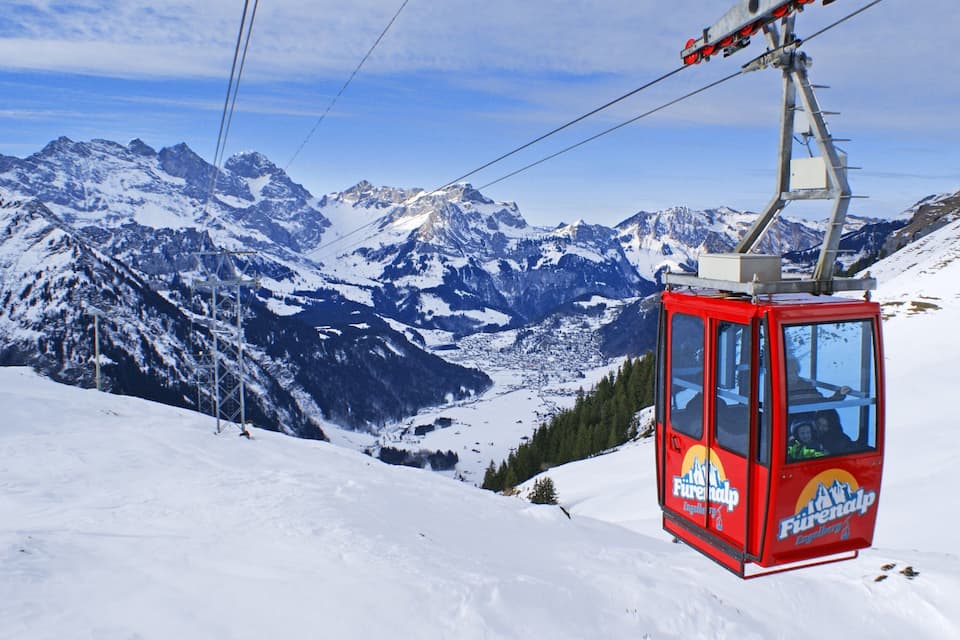 A red cable car with 'Fürenalp Engellbuh' written on it, traveling over a snowy mountain with a backdrop of snow-covered peaks and a clear sky.