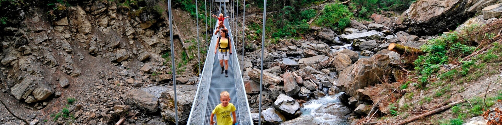 Uma ponte suspensa sobre um riacho rochoso em uma área arborizada.
