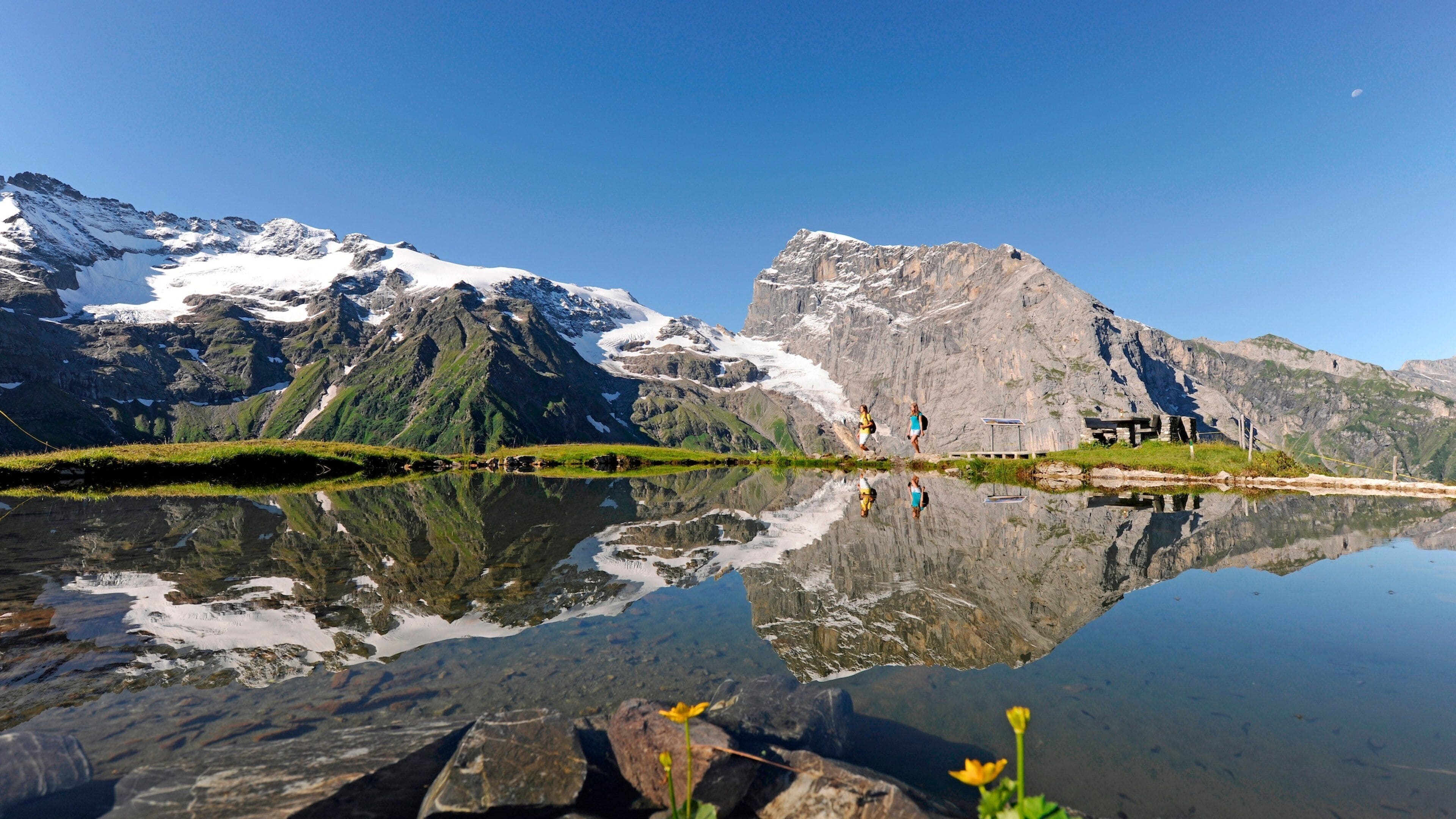 Un lago de montaña con reflejos, picos nevados y una pequeña construcción de madera.