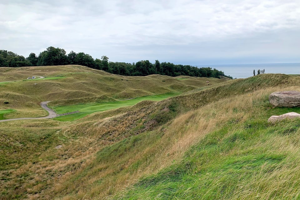 A hilly landscape with a winding road, grassy areas, and a body of water in the distance.