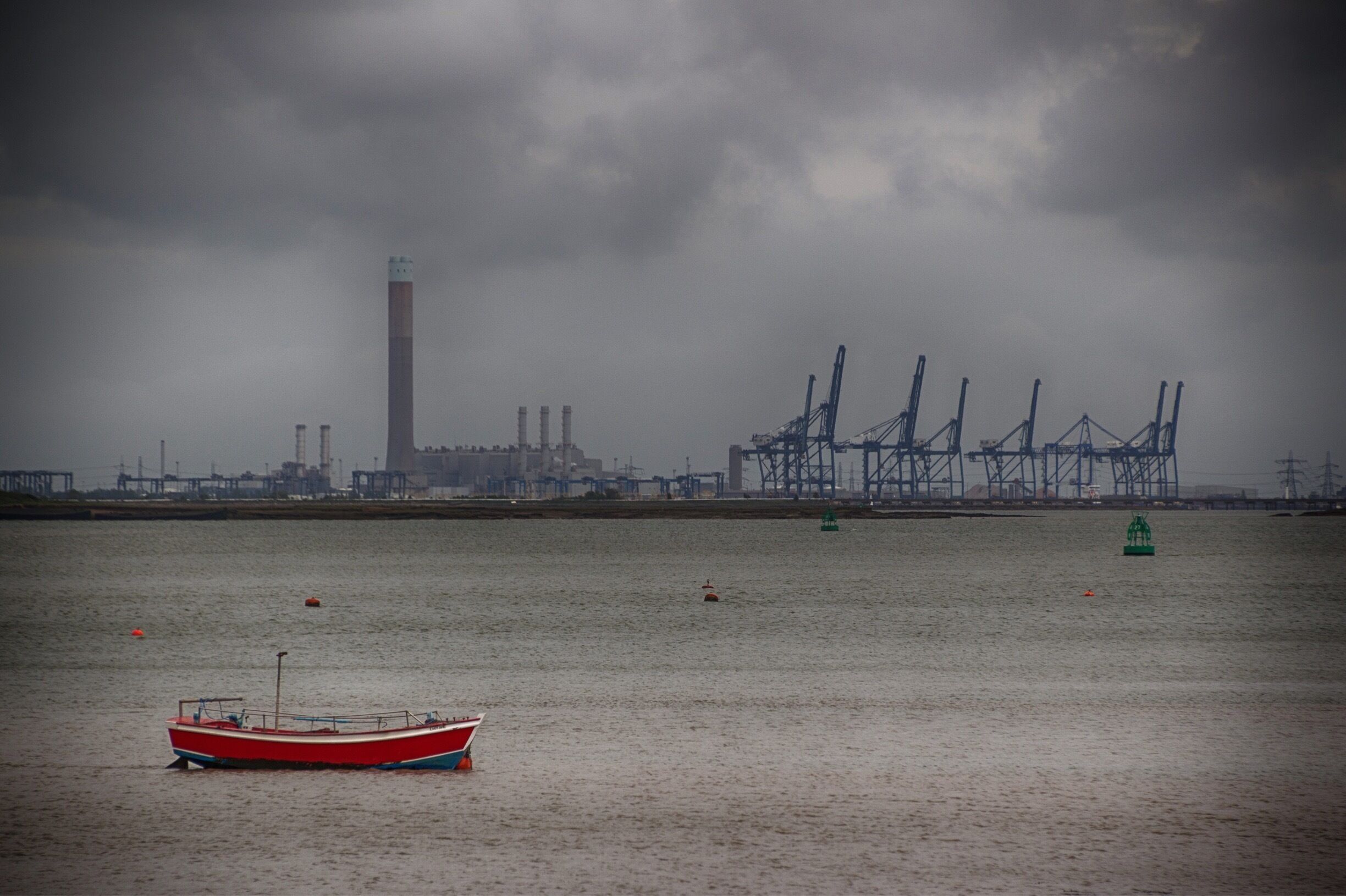 A red boat on calm water with industrial structures and a tall tower in the background.