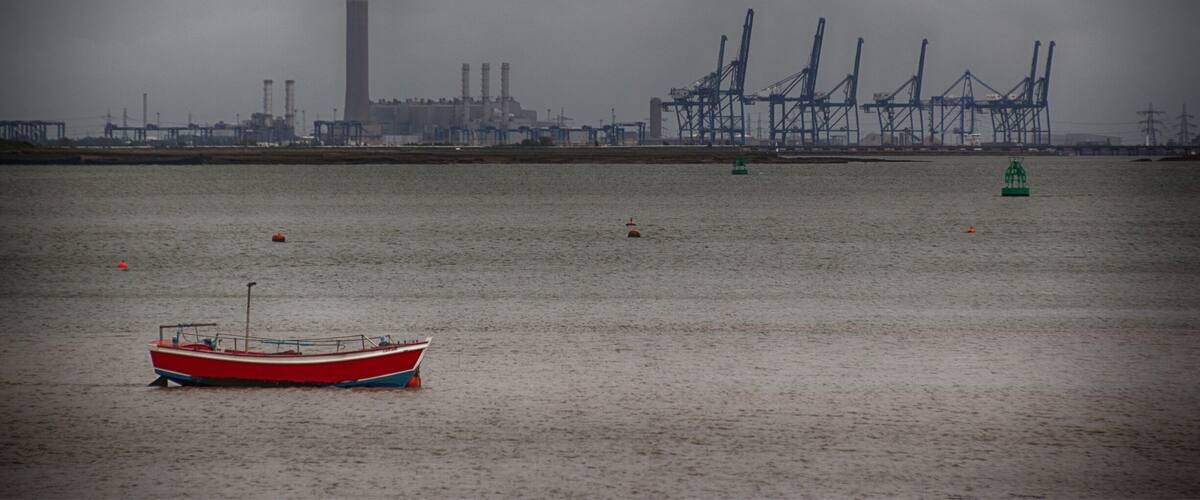 A red boat on calm water with industrial structures and a tall tower in the background.