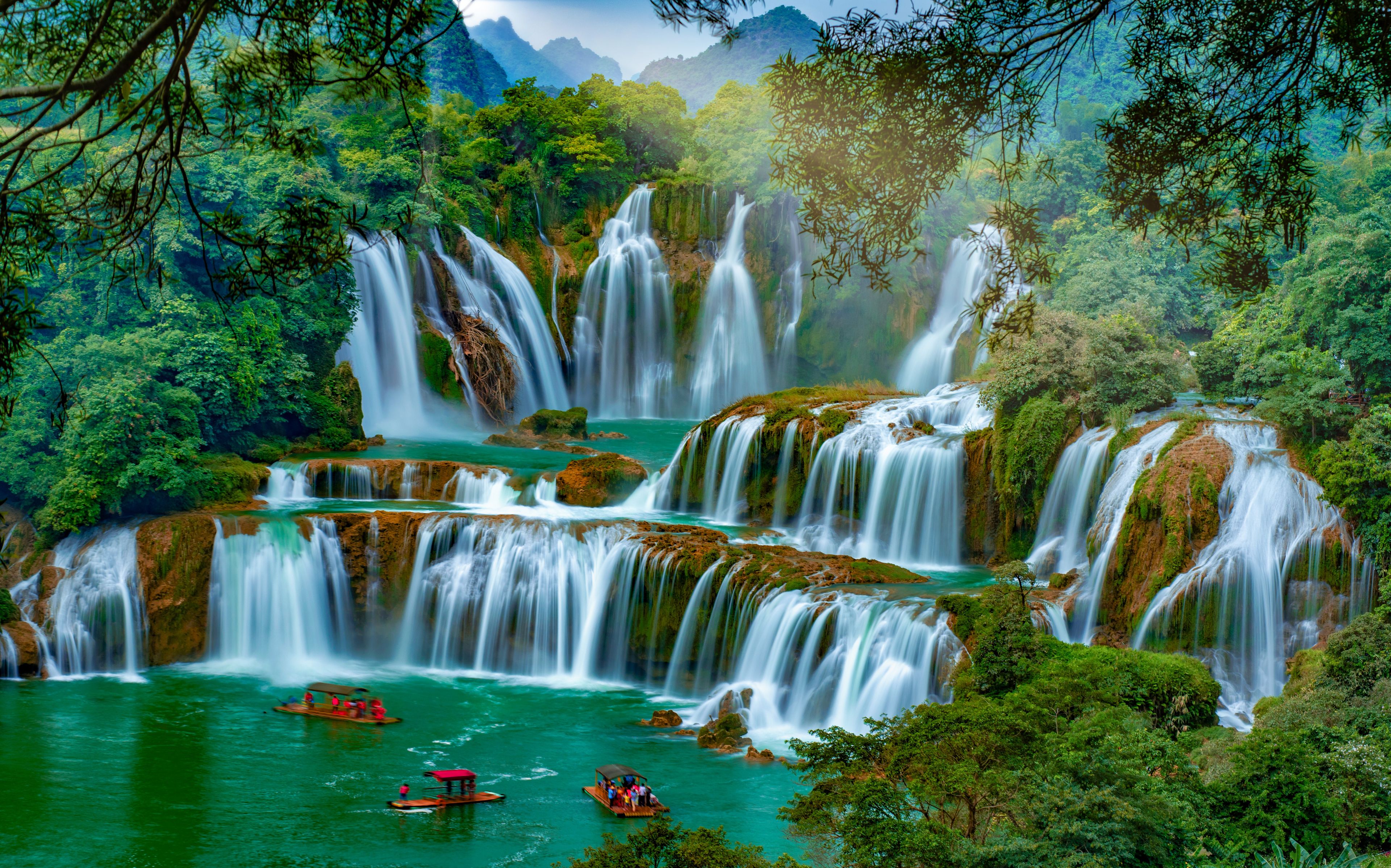 A multi-tiered waterfall surrounded by lush greenery and a river with boats.