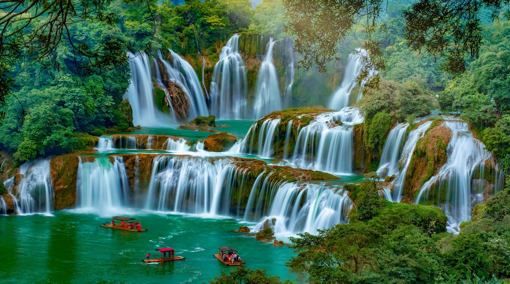 A multi-tiered waterfall surrounded by lush greenery and a river with boats.