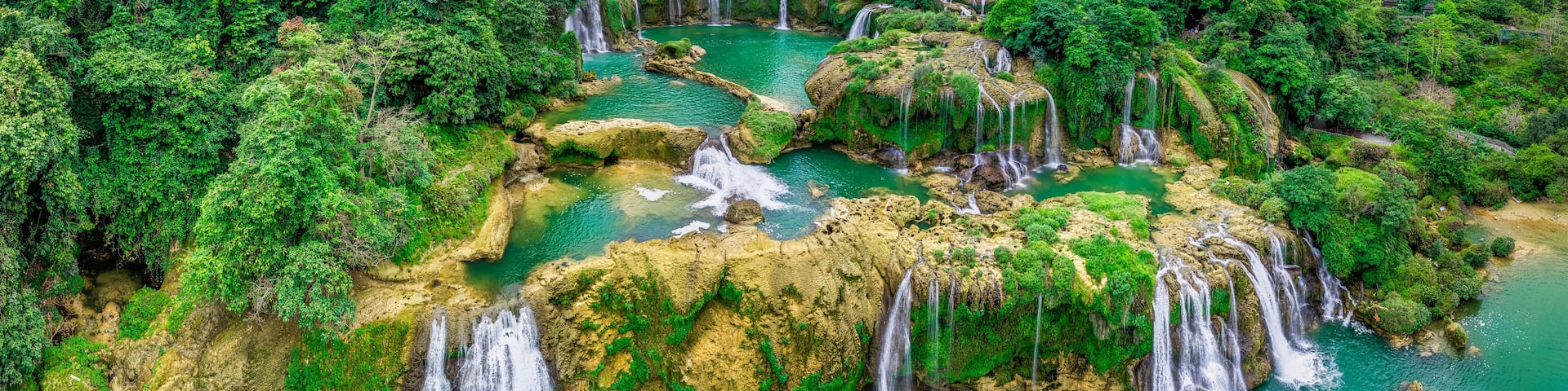A series of waterfalls cascading into a turquoise pool surrounded by lush green forest and rocky terrain.