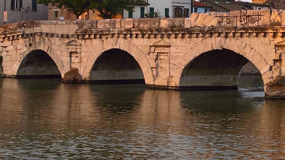 Un pont en pierre, composé de trois arches, enjambe un cours d’eau, avec des bâtiments visibles à l’arrière-plan.