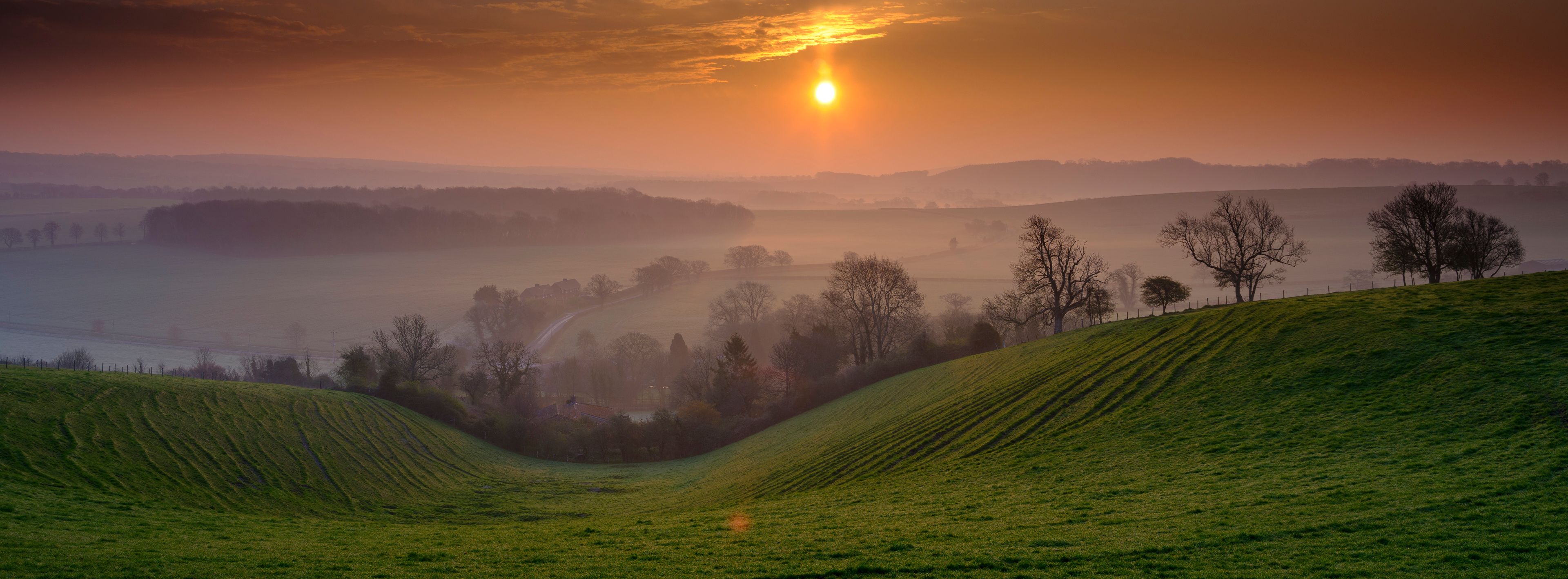 A sunrise over rolling green hills with a misty valley and scattered trees.