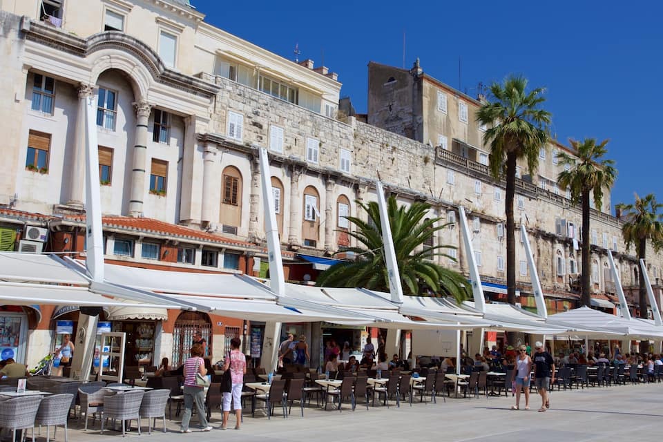 Una passeggiata lungomare con caffè all'aperto, palme e edifici storici.