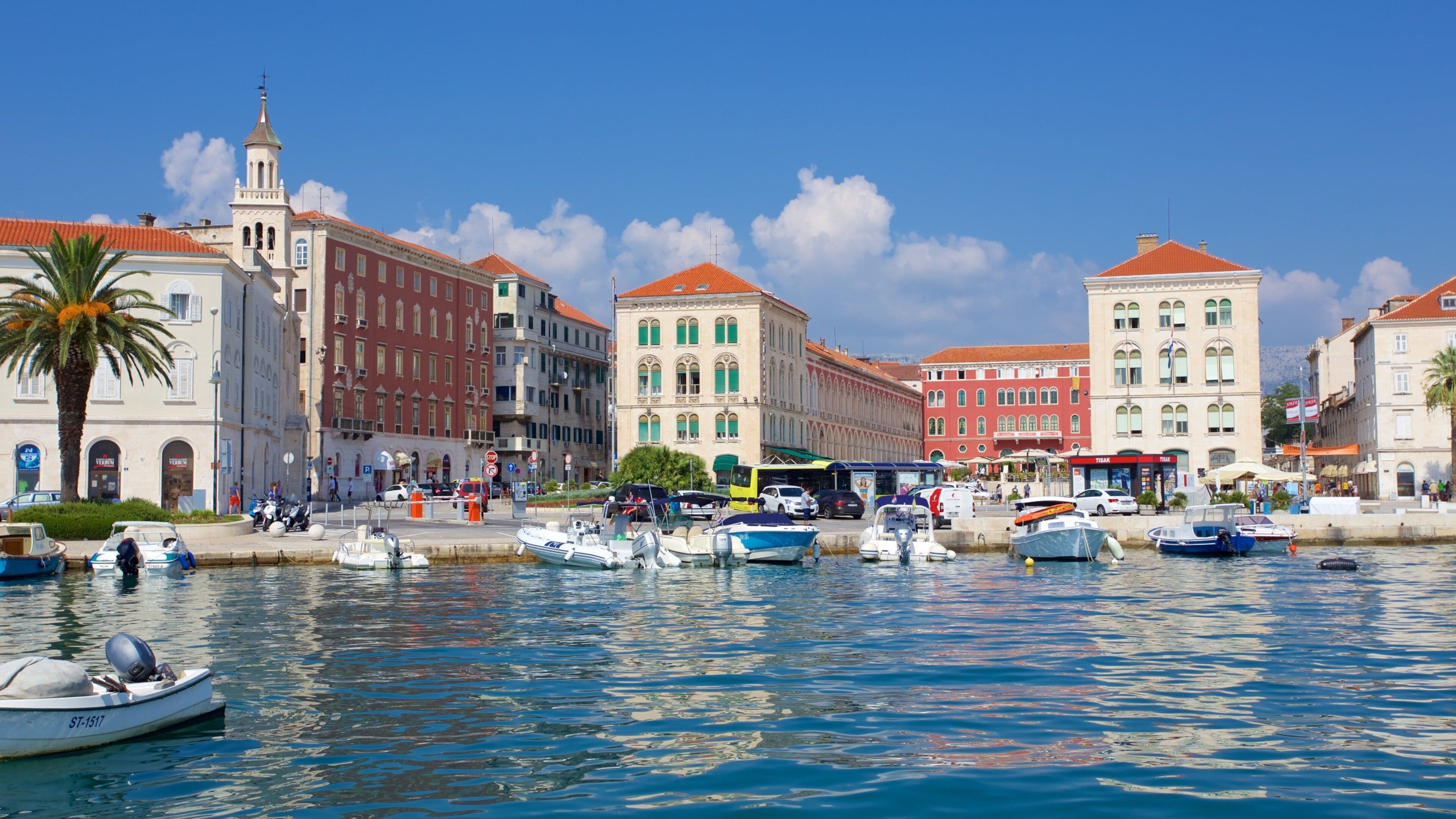 Un puerto deportivo con barcos, una palmera y edificios históricos con tejados rojos.