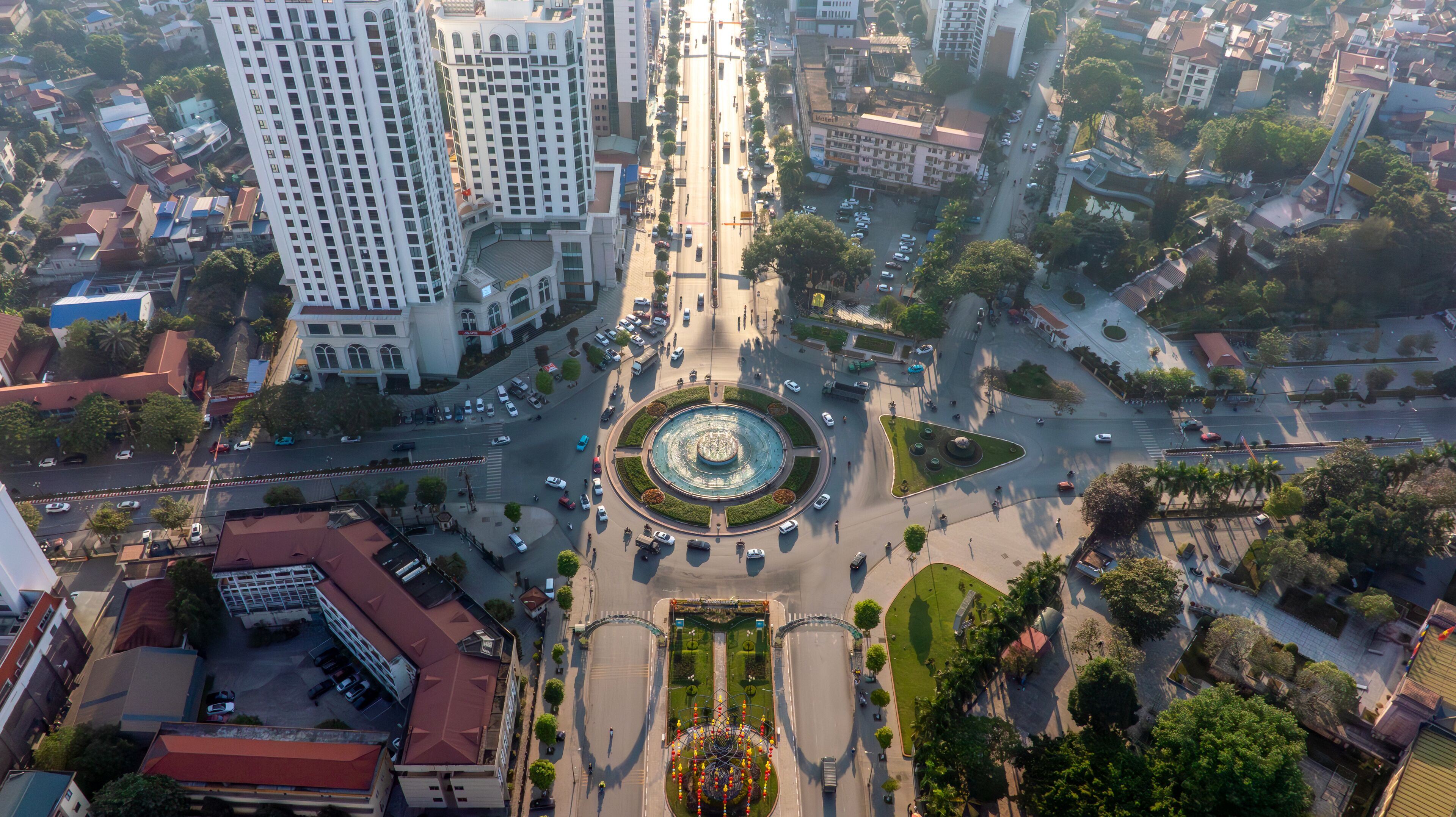 Backlit aerial view of Thai Nguyen city's central roundabout, with long shadows and sun glare, showcasing urban life under bright sunlight.