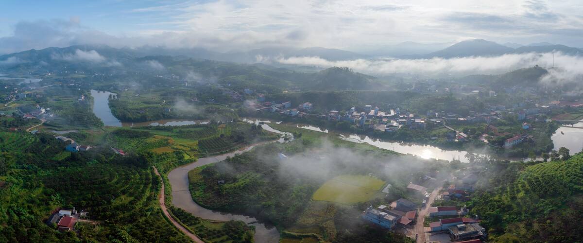 A river winding through a lush valley with scattered houses and fields, surrounded by mountains and shrouded in mist.