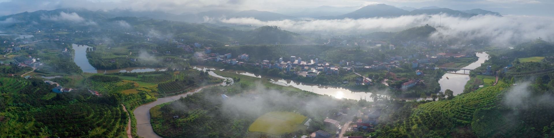 A river winding through a lush valley with scattered houses and fields, surrounded by mountains and shrouded in mist.