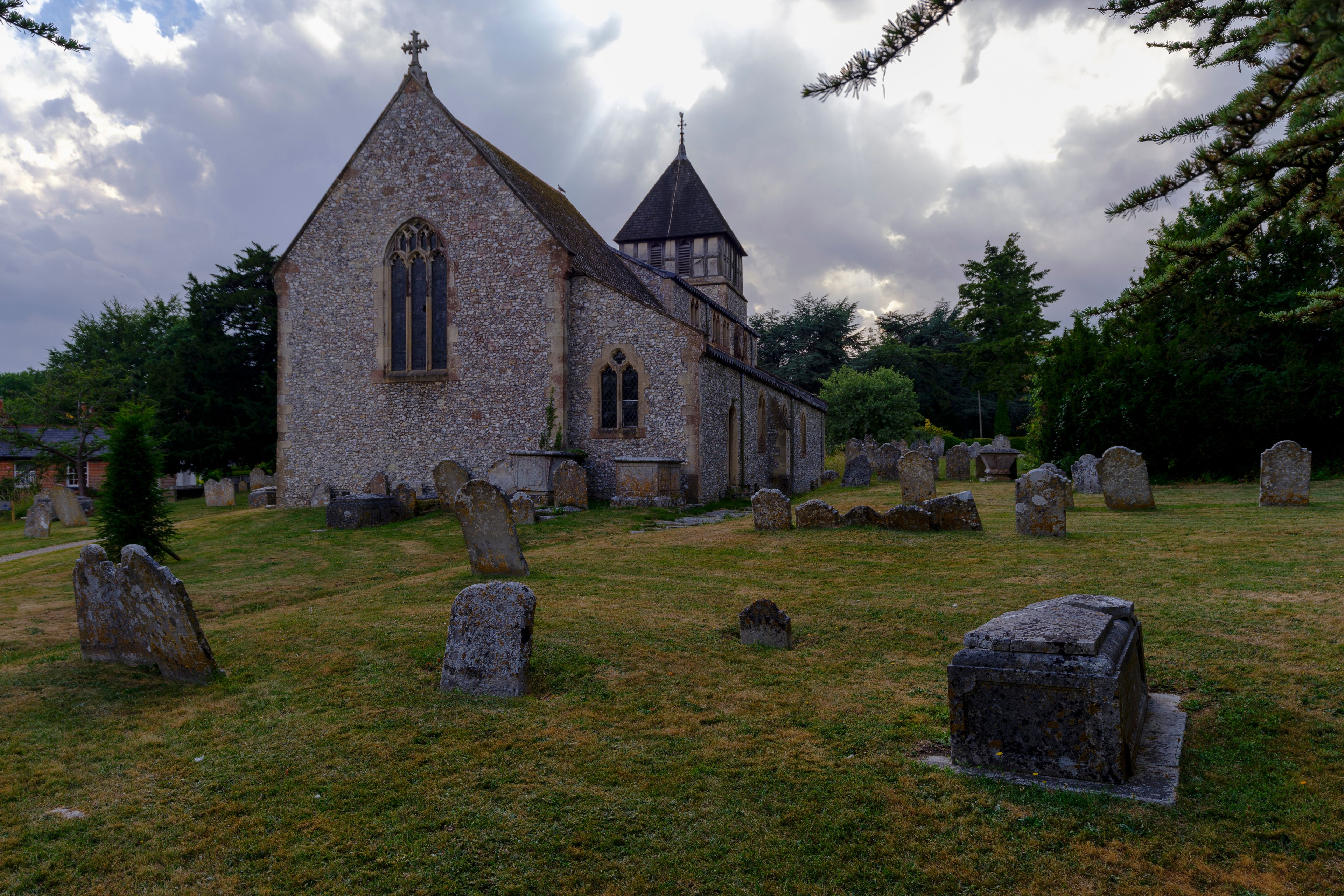 A stone church with a pointed roof and a cross on top, surrounded by a cemetery with numerous headstones.