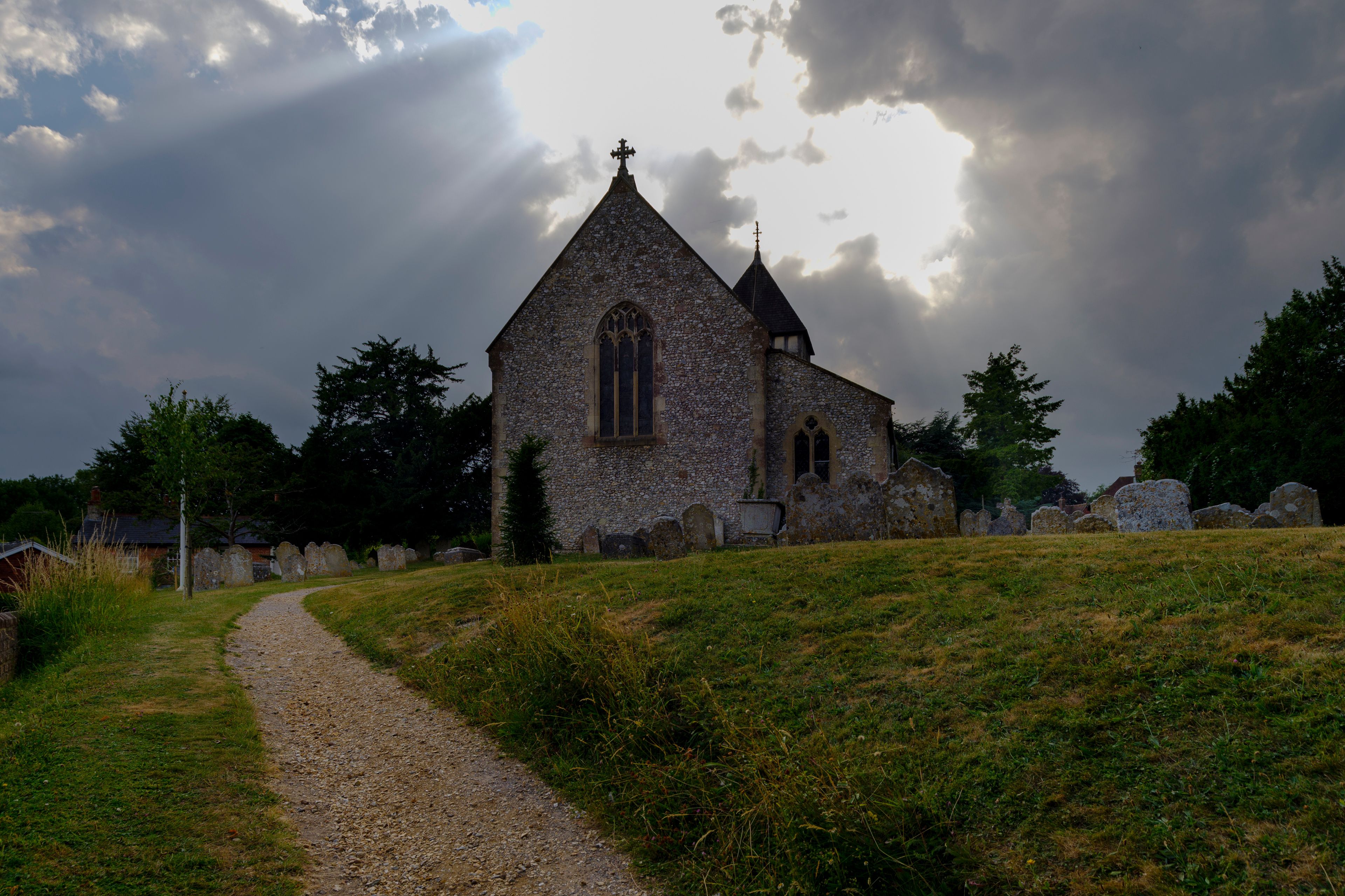 A stone church with a pointed roof and a cross on top, surrounded by a grassy area and a path leading to it.