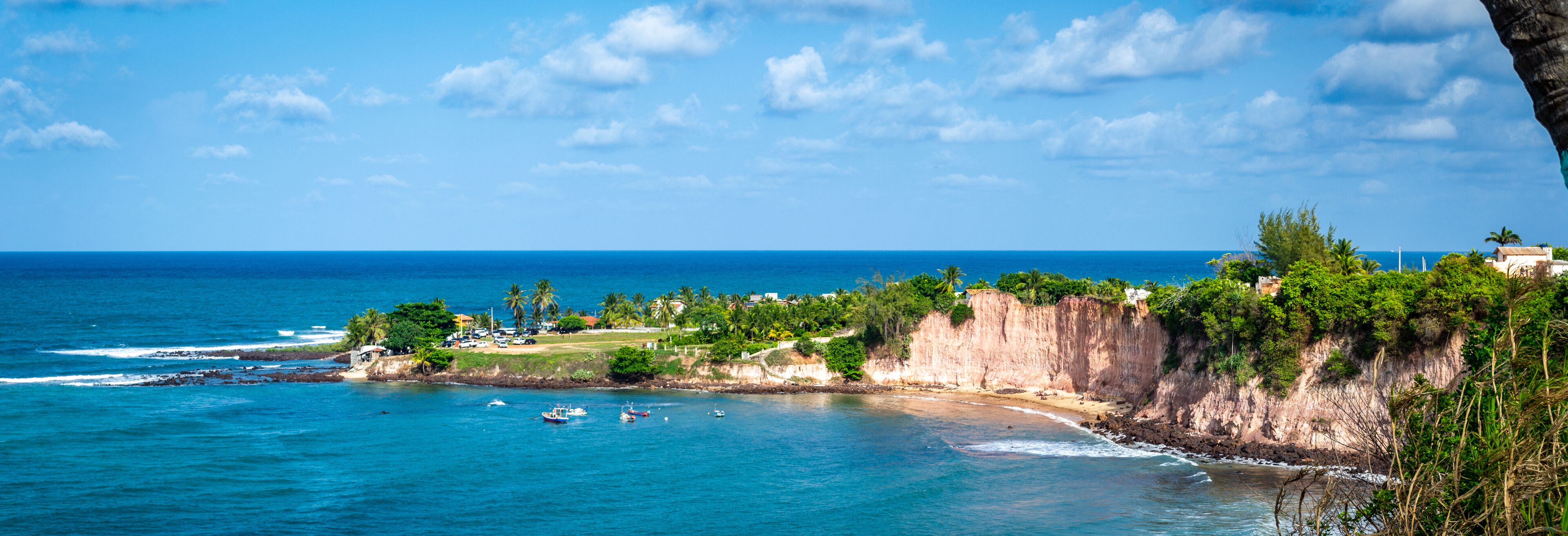 A coastal landscape with a clear blue sea, rocky cliffs, and a small town with buildings and greenery.