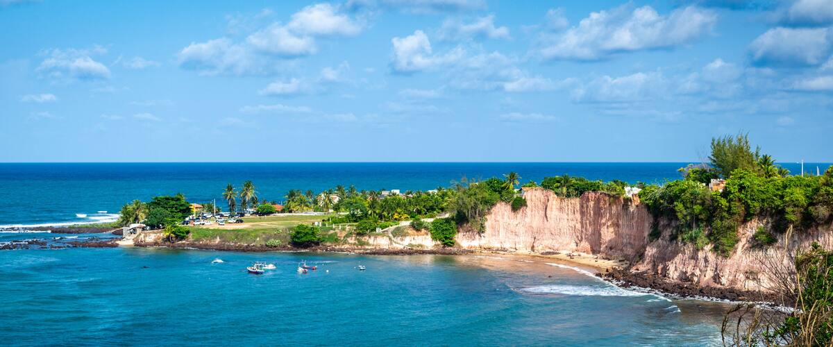 A coastal landscape with a clear blue sea, rocky cliffs, and a small town with buildings and greenery.