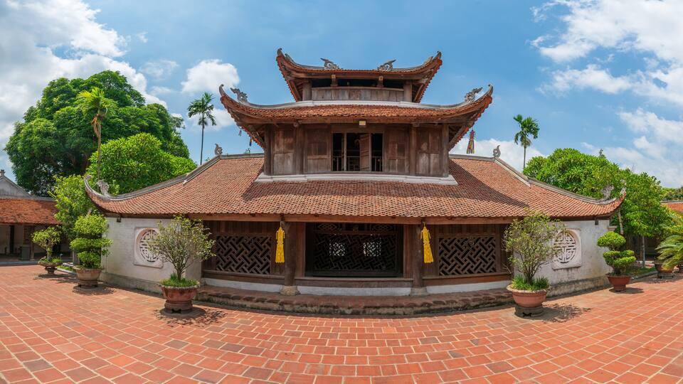 A traditional wooden building with a tiled roof and ornate decorations, surrounded by lush greenery and a paved courtyard.